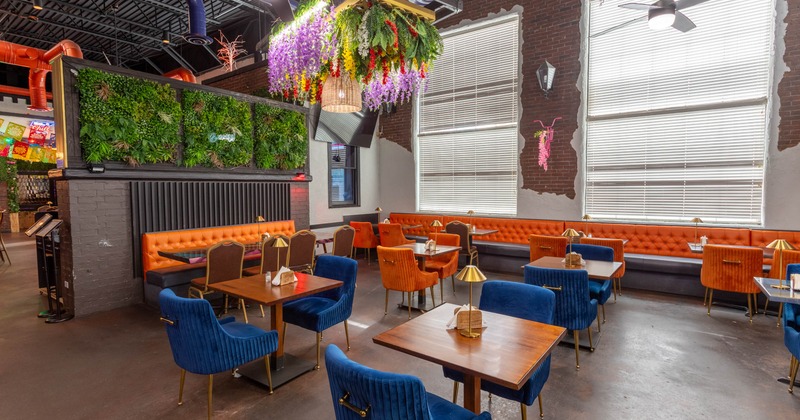 A modern restaurant interior with tufted booths, blue and orange chairs, a vertical garden, and decorative hanging flowers