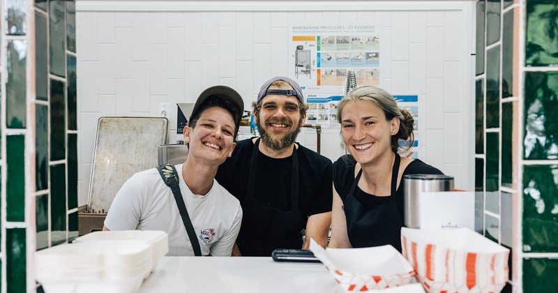Three smiling employees wearing aprons stand behind a counter in a  kitchen