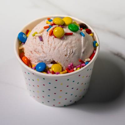 Scoop of ice cream in a paper cup topped with colorful chocolate candies and sprinkles.
