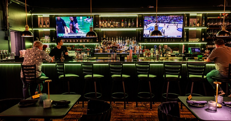 Front view to bar, bartender, drink rack, tv screens above, stools in front