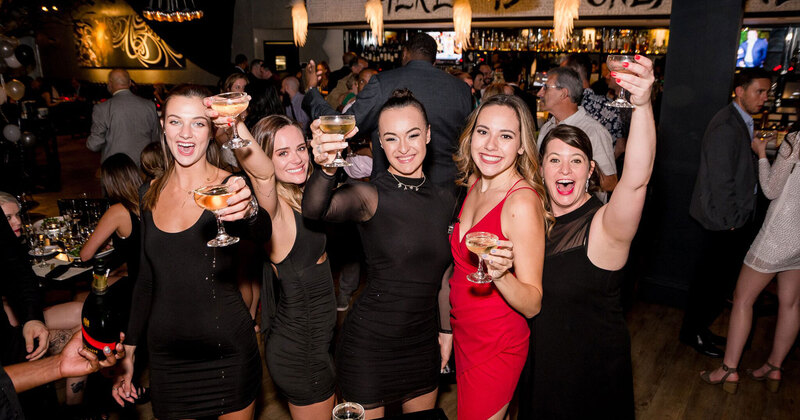 Bar area, smiling guests raise glasses as they pose for the photo, crowd of people in the background