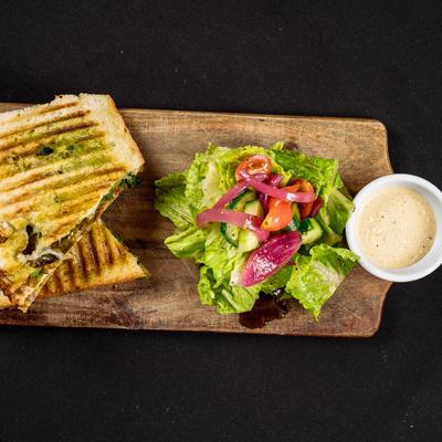 Pesto grilled cheese sandwich on a wooden board with a side salad and a cup of dressing.