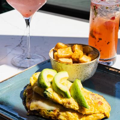 Omelet topped with avocado slices, served with a side of roasted potatoes and two drinks.