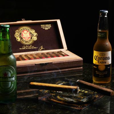 Open cigar box beside two beer bottles and two cigars on an ashtray.