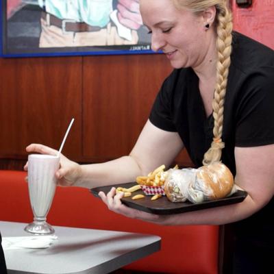 A waitperson serves a milkshake and food to a customer at a table inside.
