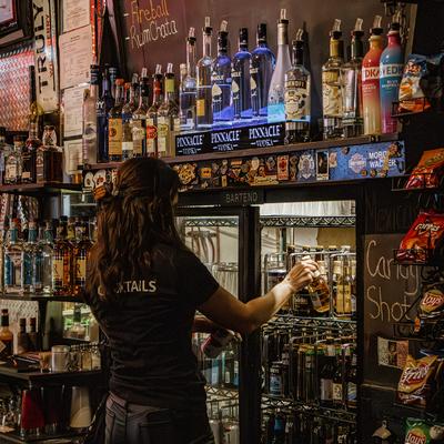 Bartender taking a beer from a beer fridge, bottles of hard liquor.