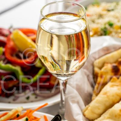 A glass of white wine on a table with food plates.
