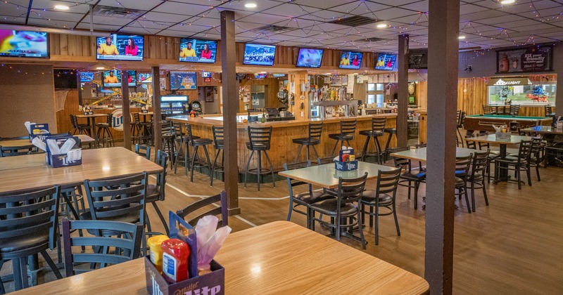 Interior of a bar with wooden tables, chairs, a central bar with stools, and TVs on the wall