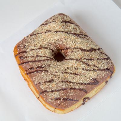 A square donut with chocolate icing and crushed nuts, drizzled with chocolate.