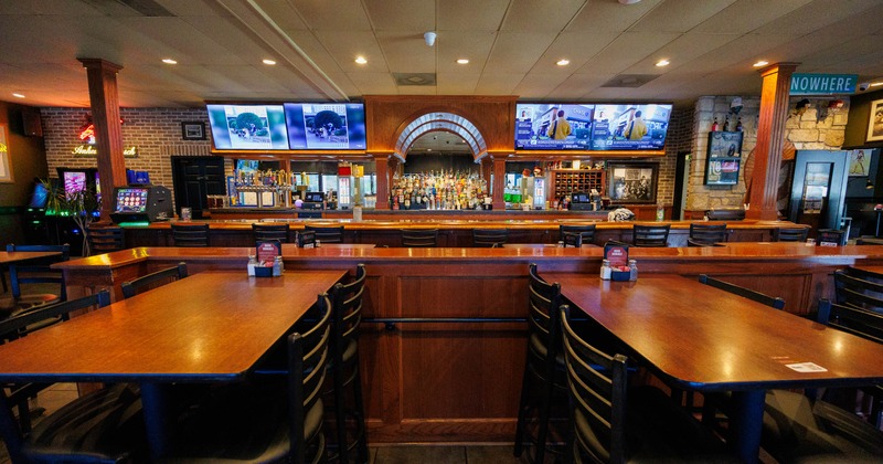 Spacious bar interior with wooden tables, chairs, a long bar counter, TV screens