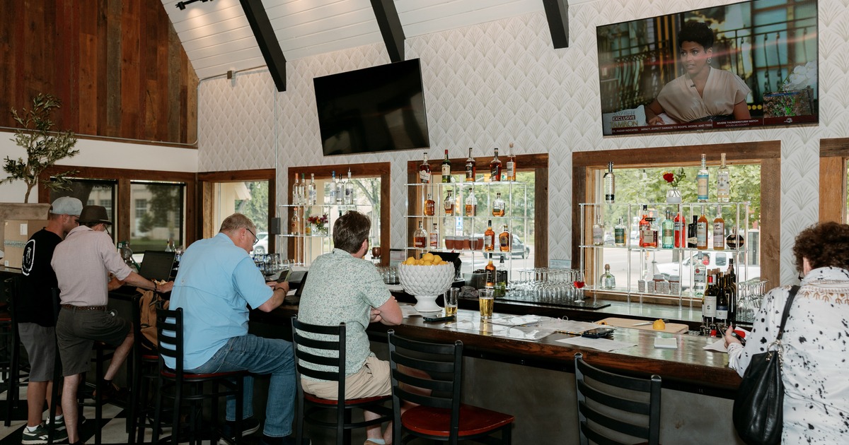 Guests seated at a bar with liquor shelves and two TVs
