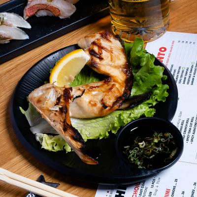 Yellowtail Collar on lettuce, with lemon, and dipping sauce on a black plate.