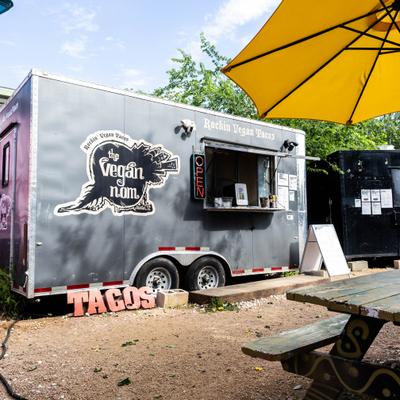 Gray food truck with a service window and signage, beside a picnic table with a umbrella.