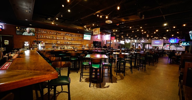 Spacious bar interior with a long wooden counter, high tables, and chairs