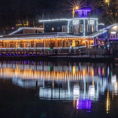 The restaurant building adorned with white and purple lights reflects on calm water at night.