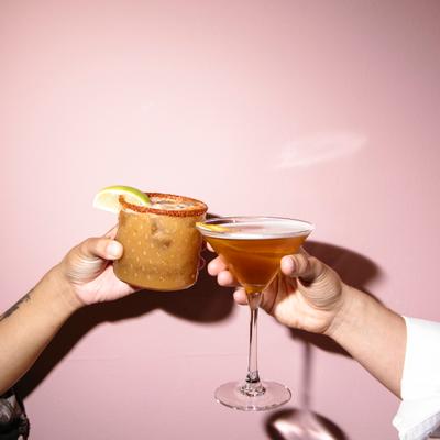 Two hands clinking cocktails against a pink wall.