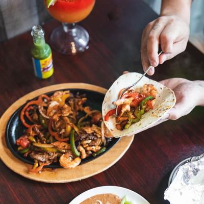 Hands assembling a shrimp and pepper fajita taco from a sizzling cast iron skillet.