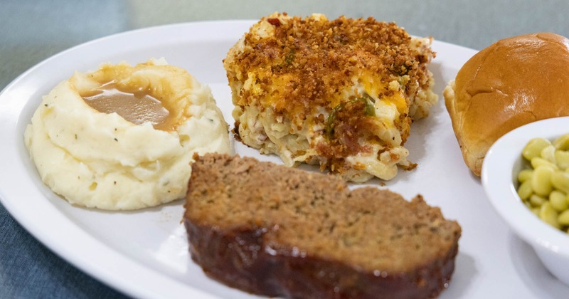 A plate with mashed potatoes and gravy, meatloaf, creamy casserole, a bun, and green beans