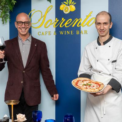 Two people posing with a glass of wine and whole pizza.