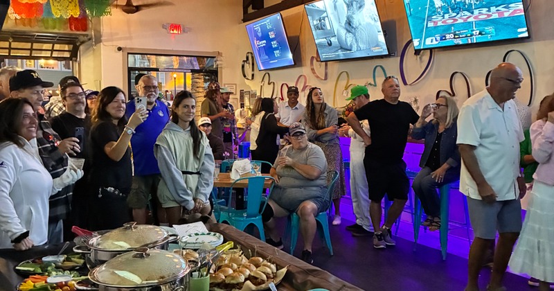 A large group of people celebrating someone out of frame with catered food and cookies
