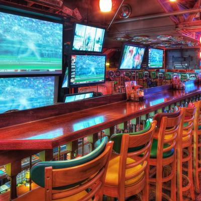 A vibrant sports bar with a long wooden counter, colorful padded bar stools, and several TVs