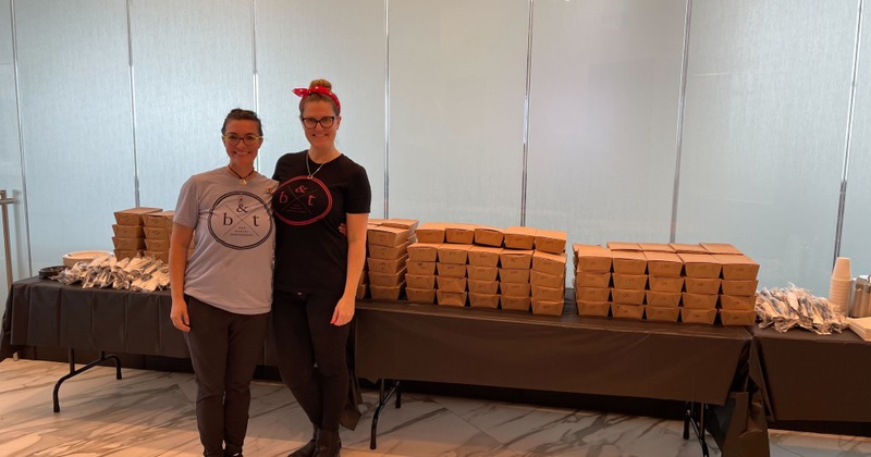Two staff members posing in front of a table with catering food packages and utensils