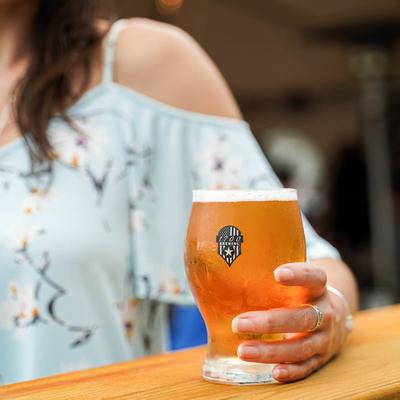 A person holds a glass of beer at an  outdoor table, the glass has the 1700 Brewing logotype.