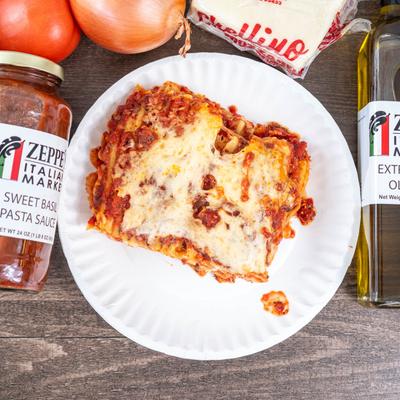A slice of lasagna on a paper plate surrounded by tomatoes, olive oil, and pasta sauce.