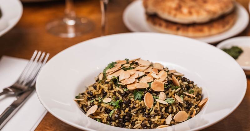 A white bowl filled with rice, lentils, and topped with sliced almonds and herbs sits on a wooden table