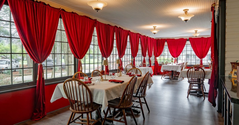 A dining room with red curtains, white tablecloths, wooden chairs and large windows