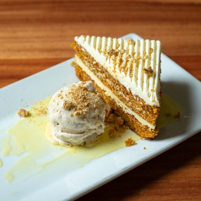A slice of carrot cake with white frosting beside a scoop of vanilla ice cream on a white plate