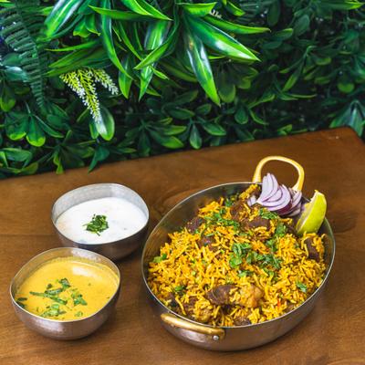 Indian lamb biryani with herbs, raita, and curry sauce served in brass bowls.