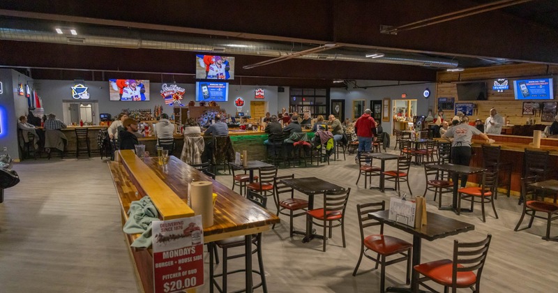 Interior of a busy bar with patrons, tables, and a long wooden bar with TVs overhead