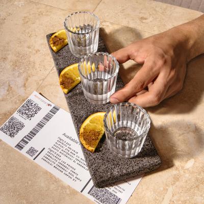 A hand reaching for a mezcal shot from a stone tray with orange slices.