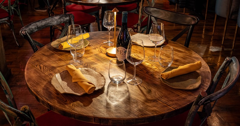Round wooden restaurant table set with yellow napkins, glasses and a wine bottle under warm lighting