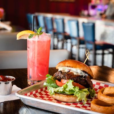 The Big Blue burger with onion rings and a glass of pink cocktail drink on a table.