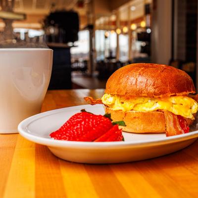 Egg and bacon sandwich on a bun served with sliced strawberries and a coffee.
