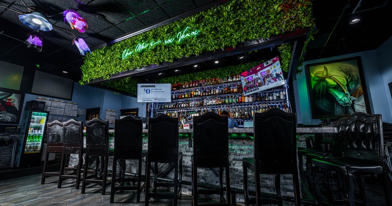 A modern bar with a green plant ceiling and neon sign above a stocked liquor shelf