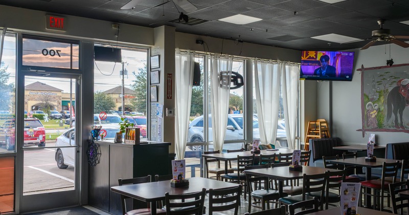 Interior of a restaurant with tables, chairs, large windows, and a TV on the wall