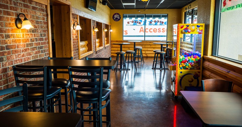 Interior of a warmly lit with brick walls, black tables and chairs, bar-height seating, and a colorful claw machine