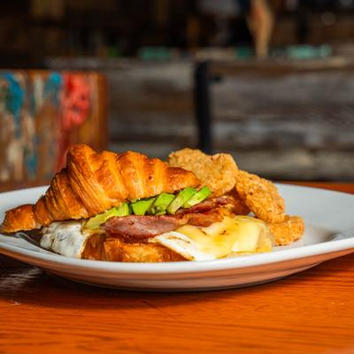 Croissant sandwich with bacon, eggs, avocado, and potato fritters.