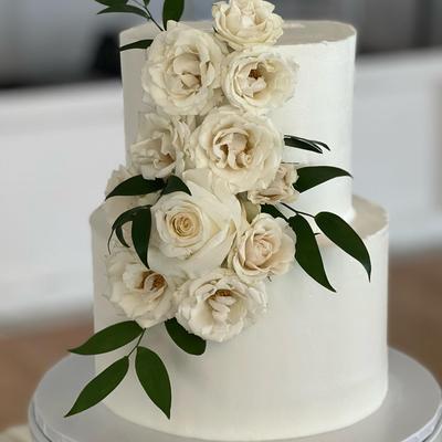 A two-tiered white wedding cake decorated with white roses and green foliage.