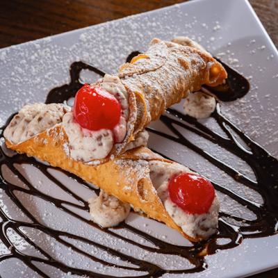 Two cannoli stuffed with ricotta cream and cherries, served on a chocolate drizzled plate.