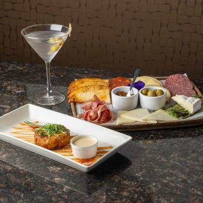 A cocktail sits beside a charcuterie board and crab cake plate.
