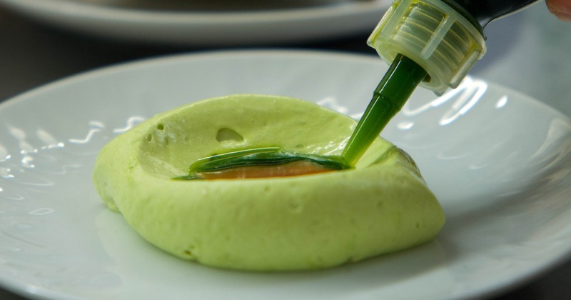 Close-up of green mousse with amber sauce and green liquid being added on a white plate