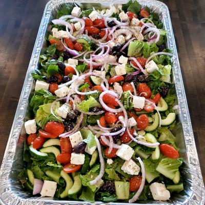Large catering tray of Greek salad topped with cubed feta cheese.