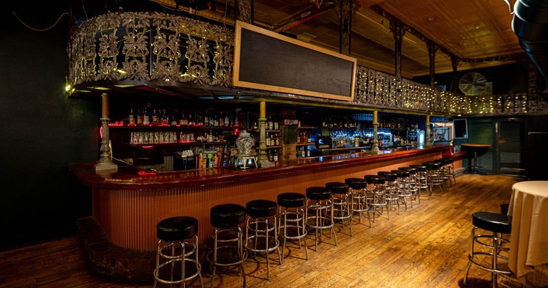 Bar interior with long wooden counter, metal stools, and decorative metalwork above