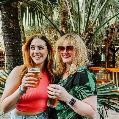 Two people posing together with beer glasses outdoors by palm trees.