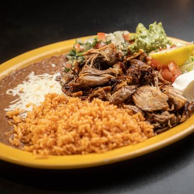 A plate of carnitas with rice, refried beans, guacamole, lettuce tomato salad, and sour cream.
