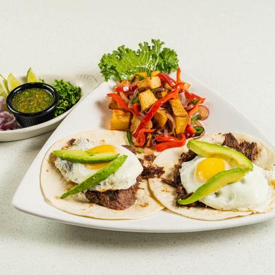 Steak tacos topped with sunny side up eggs and avocado, served with sides